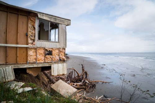 Abandoned beach house with damaged walls, debris, and a view of the ocean under a cloudy sky.