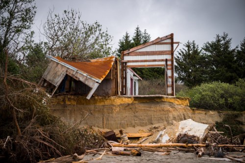 A partially collapsed house on a cliff edge, surrounded by fallen trees and debris, under a cloudy sky.