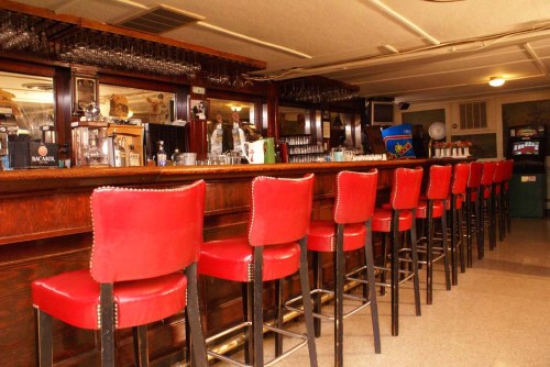 A vintage bar interior featuring a long wooden counter and red upholstered stools. Glassware is displayed on shelves.