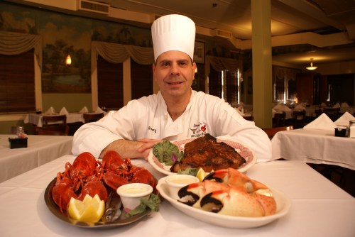 A chef in a white hat poses with a platter of seafood and grilled dishes in a restaurant setting.
