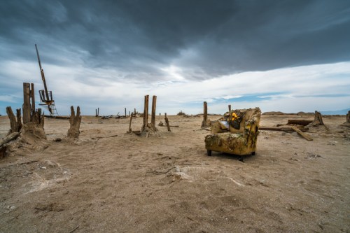 An abandoned, weathered armchair sits on a barren landscape under a cloudy sky, surrounded by remnants of old structures.