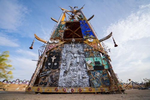 A tall, colorful structure made of various materials and objects, set against a clear blue sky.