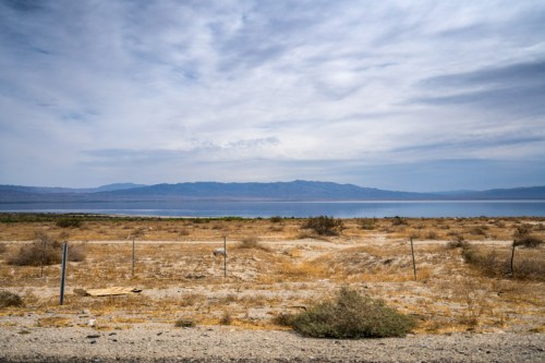A dry, barren landscape with sparse vegetation, leading to a calm body of water and distant mountains under a cloudy sky.