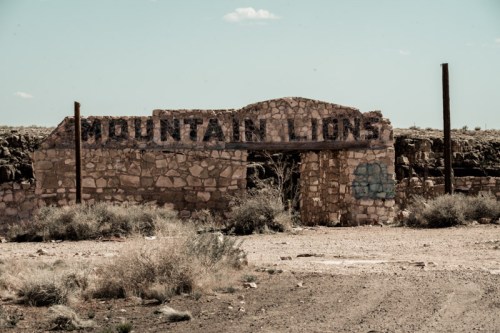 Weathered stone archway with the words "MOUNTAIN LIONS" above, surrounded by arid landscape and sparse vegetation.