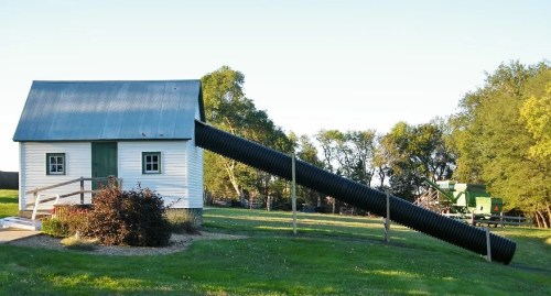 A small white barn with a metal roof, featuring a large black pipe extending from it, set in a grassy field.