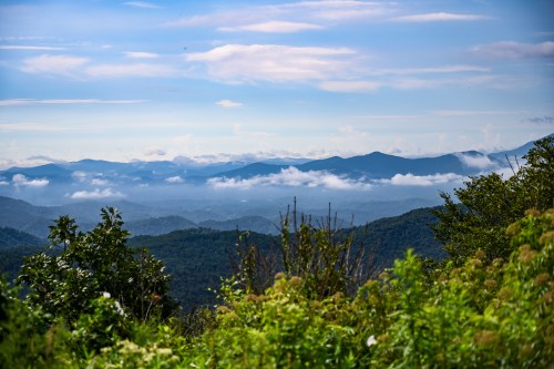 A scenic view of mountains and valleys, with clouds hovering over lush greenery under a clear blue sky.