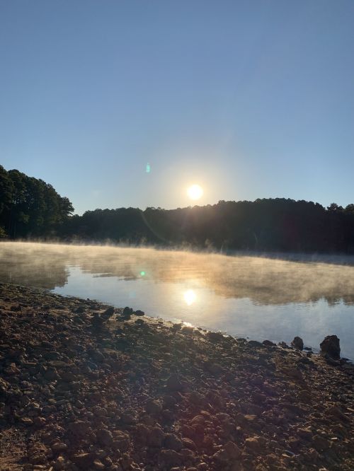 A serene lake at sunrise, with mist rising from the water and trees lining the shore.