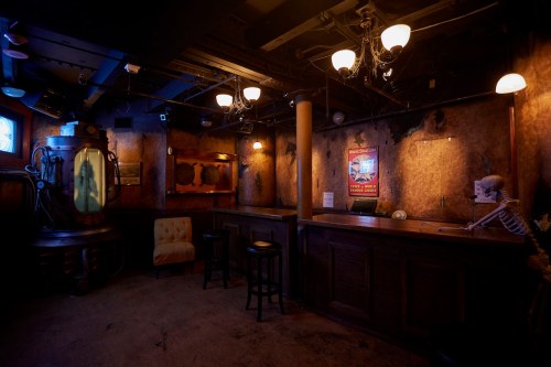 A dimly lit bar with vintage decor, featuring a skeleton, stools, and a large glass tank.