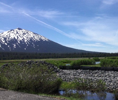A snow-capped mountain rises above a green landscape with a clear blue sky and a winding river in the foreground.