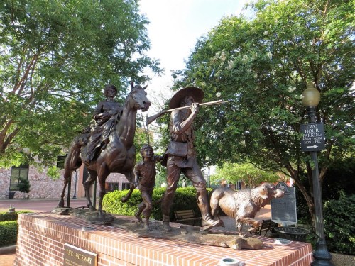 Bronze statue depicting a cowboy on horseback, a boy, and a dog, surrounded by greenery and a parking sign.