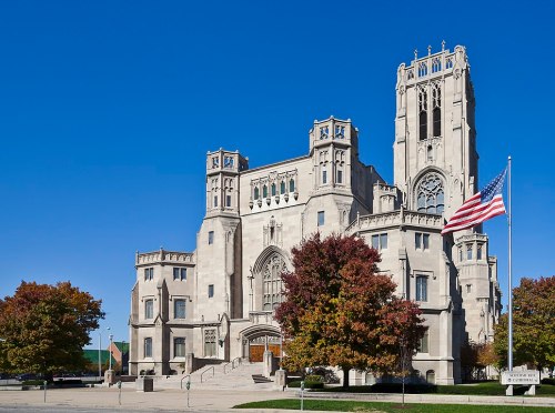 A large, ornate stone building with a tall tower, surrounded by colorful trees and an American flag in front.