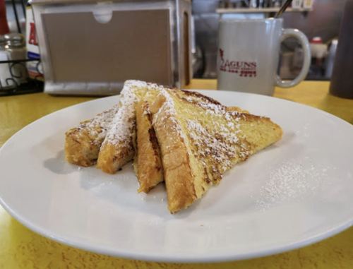 Three slices of French toast dusted with powdered sugar on a white plate, with a coffee mug in the background.