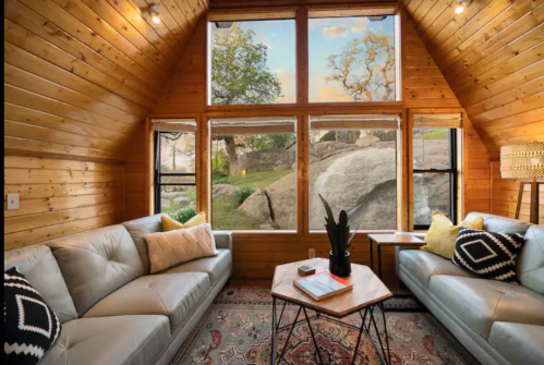 Cozy cabin interior with wooden walls, large windows, and a view of rocks and trees outside. Stylish furniture and decor.