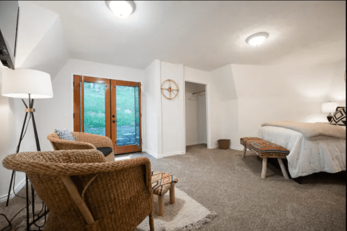 Cozy bedroom with two chairs, a bed, and a door leading to a green outdoor area. Soft lighting and neutral decor.