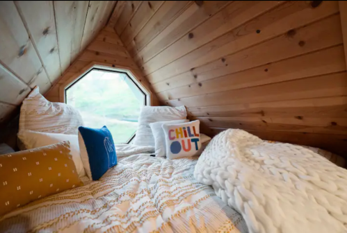 Cozy wooden loft with a bed, decorative pillows, and an octagonal window offering a serene view.