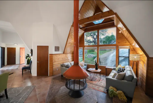 Cozy interior of a modern cabin with a triangular roof, large windows, and a central fireplace.