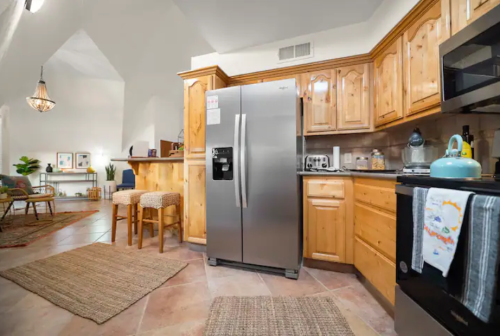 A cozy kitchen with wooden cabinets, stainless steel fridge, and a living area in the background, featuring a warm ambiance.