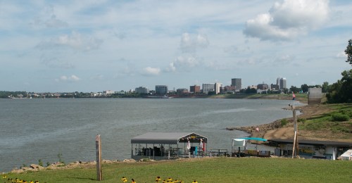 A view of a river with a city skyline in the background, featuring clouds and a sandy shore in the foreground.