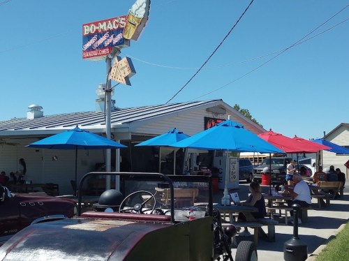 A roadside ice cream shop with colorful umbrellas, outdoor seating, and a vintage car in the foreground.