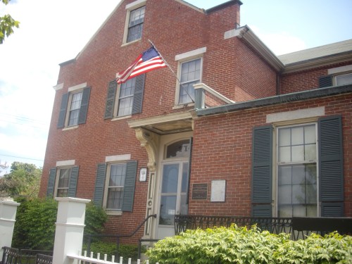 A brick building with large windows, an American flag, and green shrubs in front.