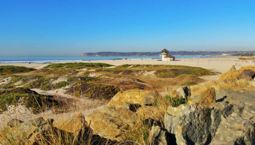 A sandy beach with gentle waves, a lifeguard tower in the distance, and rocky foreground under a clear blue sky.