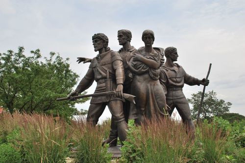 A bronze statue of four figures in historical attire, standing together in a natural setting with tall grass.