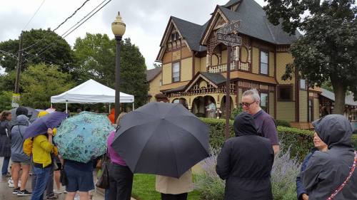 A line of people with umbrellas waiting outside a large, ornate Victorian house on a cloudy day.