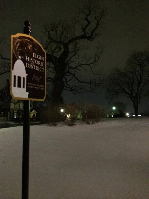 Sign for Elgin Historic District in a snowy landscape at night, with a large tree and faint lights in the background.