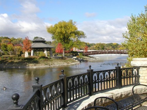 Scenic view of a park with a pond, colorful trees, a gazebo, and a wooden bridge under a partly cloudy sky.