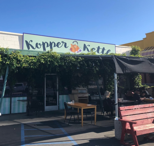 Exterior of Kopper Kettle restaurant with outdoor seating, greenery, and a clear blue sky.