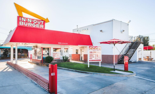 A bright red and white In-N-Out Burger restaurant with a large sign, outdoor seating, and a staircase on the side.