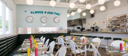 Bright restaurant interior with green seating, round lights, and a bar, featuring clocks and a cheerful wall quote.