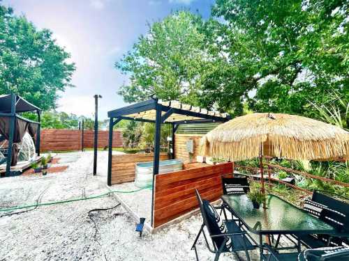 A backyard scene featuring a gazebo, a thatched umbrella, and a seating area surrounded by greenery and gravel pathways.