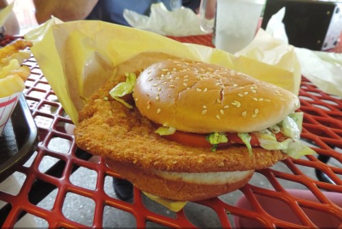 A crispy chicken sandwich with lettuce and tomato on a sesame seed bun, served on a yellow paper wrapper.