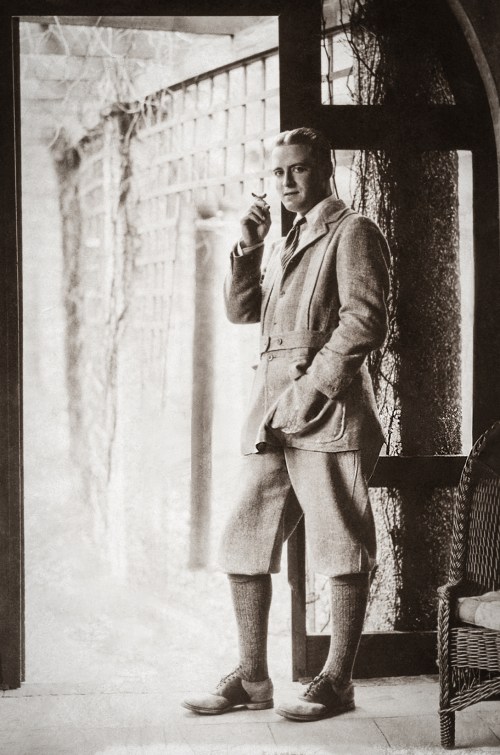 A vintage black-and-white photo of a man in a stylish outfit, standing by a doorway with a cigarette.