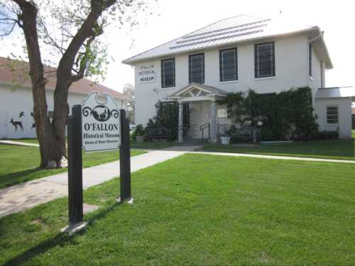 O'Fallon Historical Museum building with a sign, surrounded by green grass and trees on a sunny day.