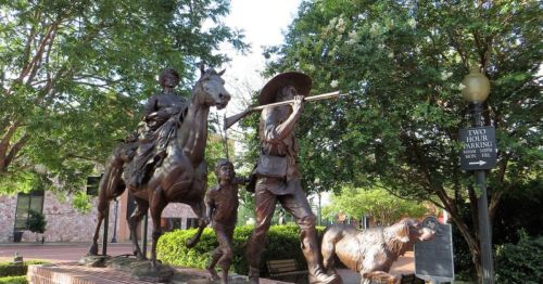 Bronze statue of a man playing a flute, accompanied by a child, a horse, and a dog, set in a green park.