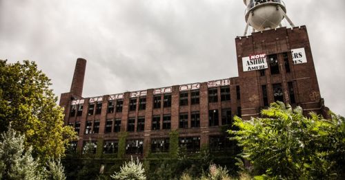 Abandoned brick factory with overgrown vegetation and a water tower, featuring graffiti and a cloudy sky.