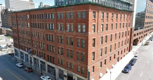 A large red brick building with multiple windows, located on a city street, surrounded by other buildings and parked cars.