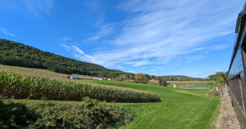 A scenic landscape featuring green fields, corn rows, and rolling hills under a bright blue sky with scattered clouds.
