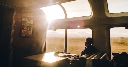 A person gazes out a train window at a sunlit landscape, with a table and notebook nearby.