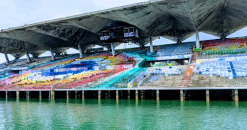 Abandoned stadium with colorful, worn seating and water reflecting beneath the structure.