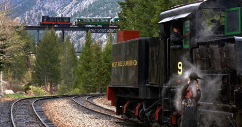 A vintage steam train passes a bridge, while a worker waves from the front of another train on a scenic railway.