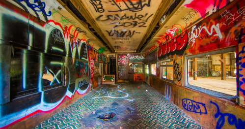 Interior of an abandoned train car covered in colorful graffiti, with a patterned floor and light streaming through windows.