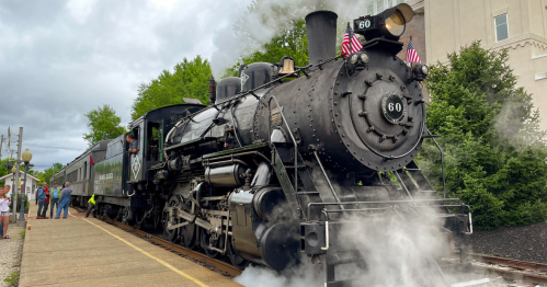 A vintage steam locomotive with smoke billowing, parked on a platform with people nearby and greenery in the background.