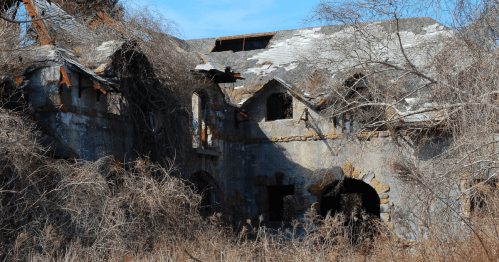 An abandoned, overgrown house with a crumbling roof and vines covering its stone walls under a clear blue sky.