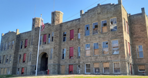 Abandoned stone building with boarded windows and an American flag, set against a clear blue sky.