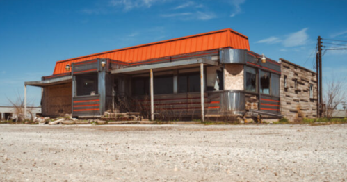 Abandoned diner with a bright orange roof, weathered exterior, and overgrown surroundings under a clear blue sky.