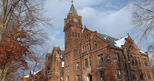 Historic brick building with a tall clock tower, surrounded by trees and a cloudy sky. Snow is visible on the roof.
