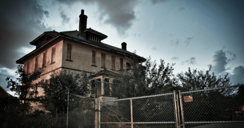 Abandoned two-story house surrounded by overgrown trees and a chain-link fence, under a moody sky.